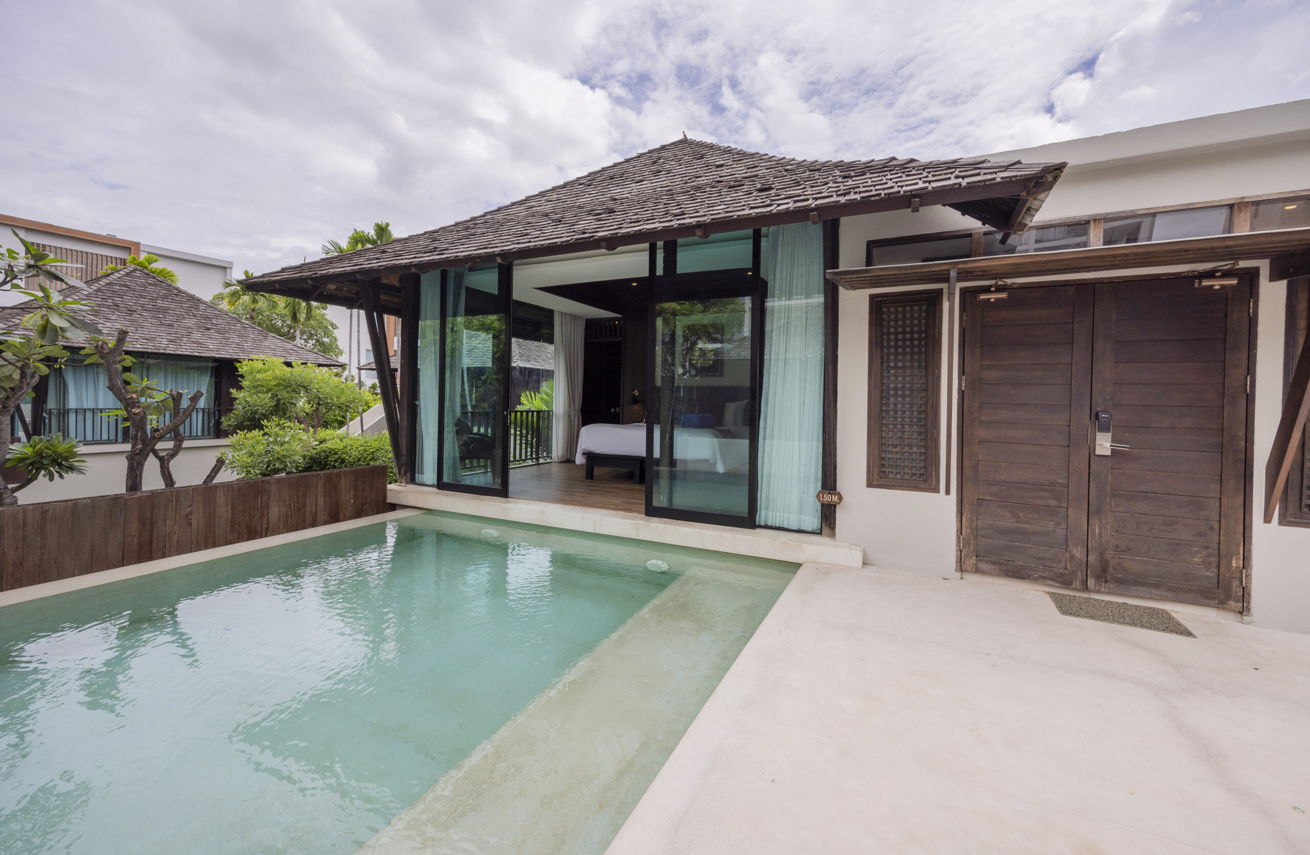 A modern villa with a private pool, glass doors leading to a bedroom, wooden roof, and cloudy sky above. Lush greenery surrounds the peaceful, tropical outdoor area.