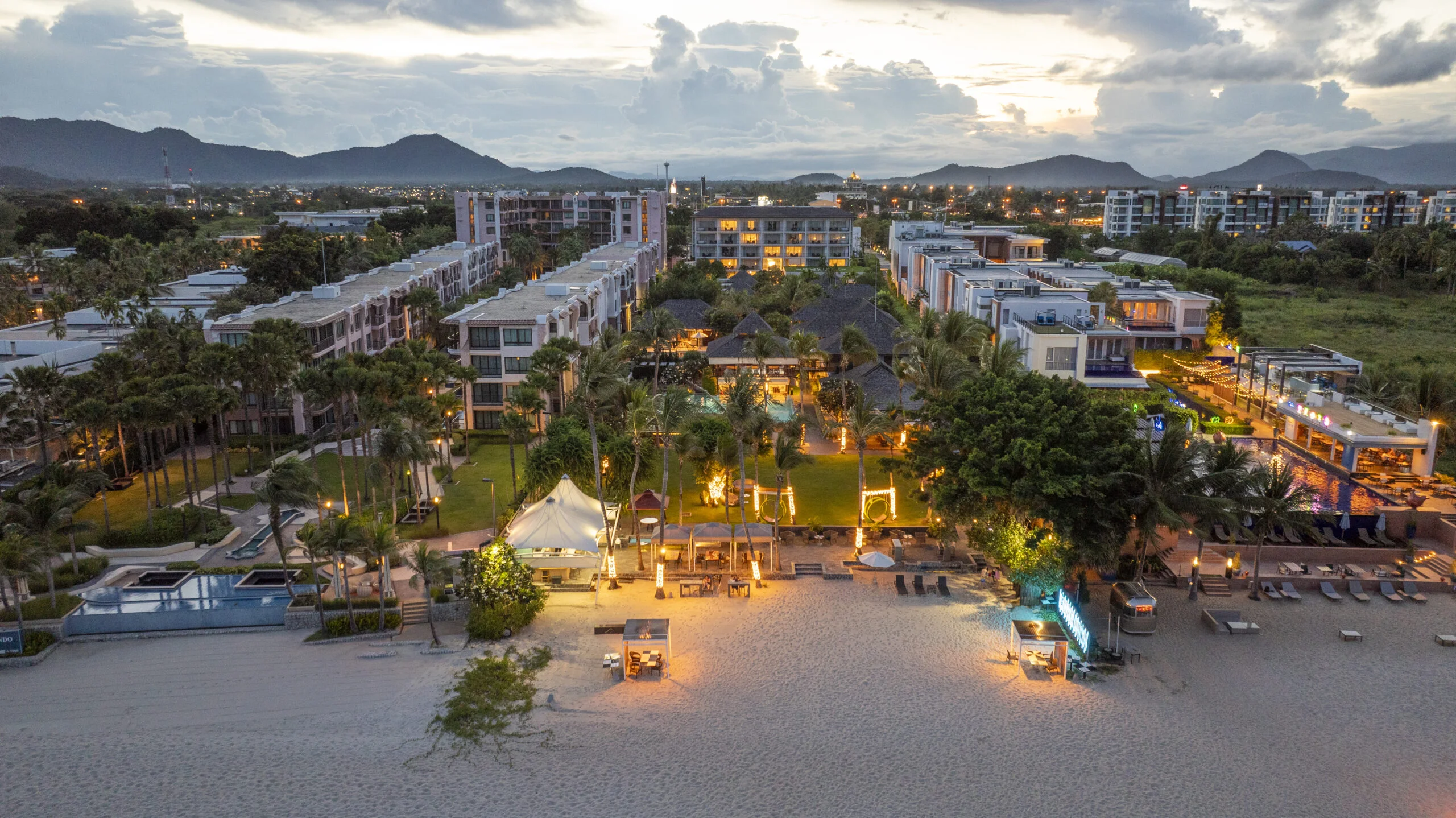 Aerial view of a luxury hotel Hua Hin beachfront resort at sunset, with illuminated pathways, modern buildings, palm trees, lounge chairs on the sand, and mountains in the background.