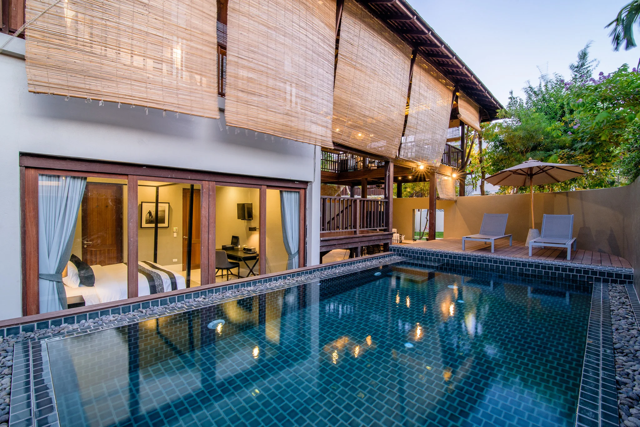 A modern pool villa in Hua Hin featuring a private tiled pool, poolside lounge chairs, large glass doors leading into the bedroom, and bamboo shades hanging from the upper floor, all surrounded by lush greenery.