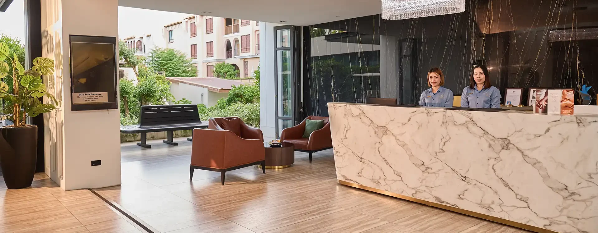 A modern hotel lobby with a marble reception desk, two staff behind it, and brown chairs nearby. Large windows reveal greenery and apartment buildings outside.