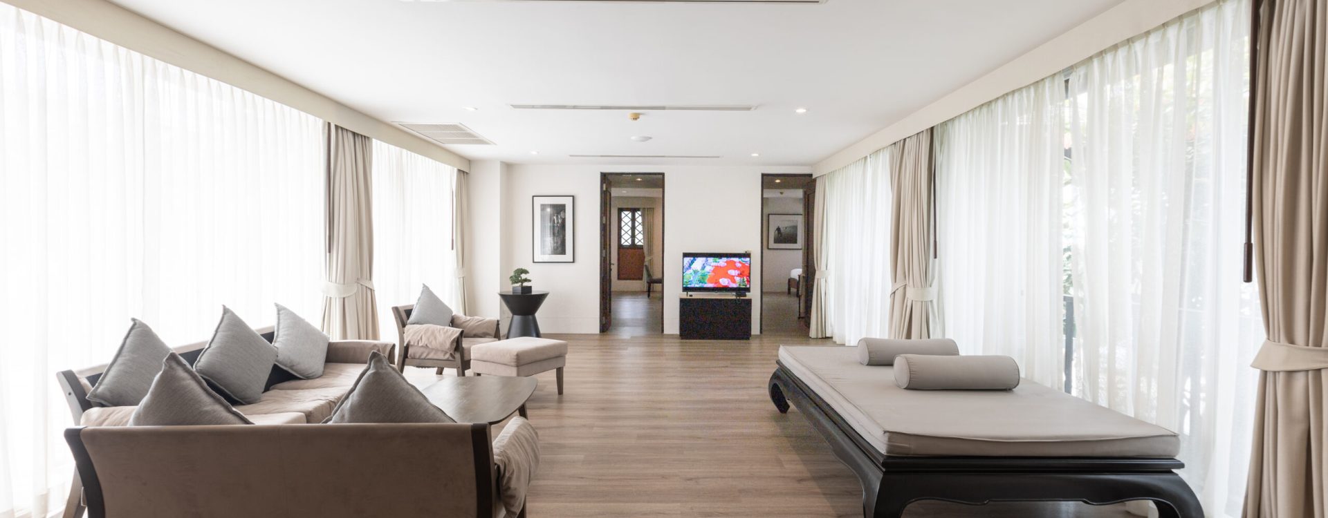 Spacious, modern living room with large windows, sheer curtains, light wood floor, a sofa set with gray cushions, a black daybed, and a TV in the far corner. Natural light fills the space.