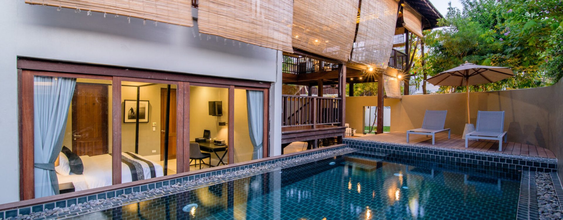 Luxury villa with a private pool, wooden deck, two lounge chairs, large windows, and bamboo shades. Indoor areas are visible through glass doors, surrounded by greenery and warm outdoor lighting at dusk.