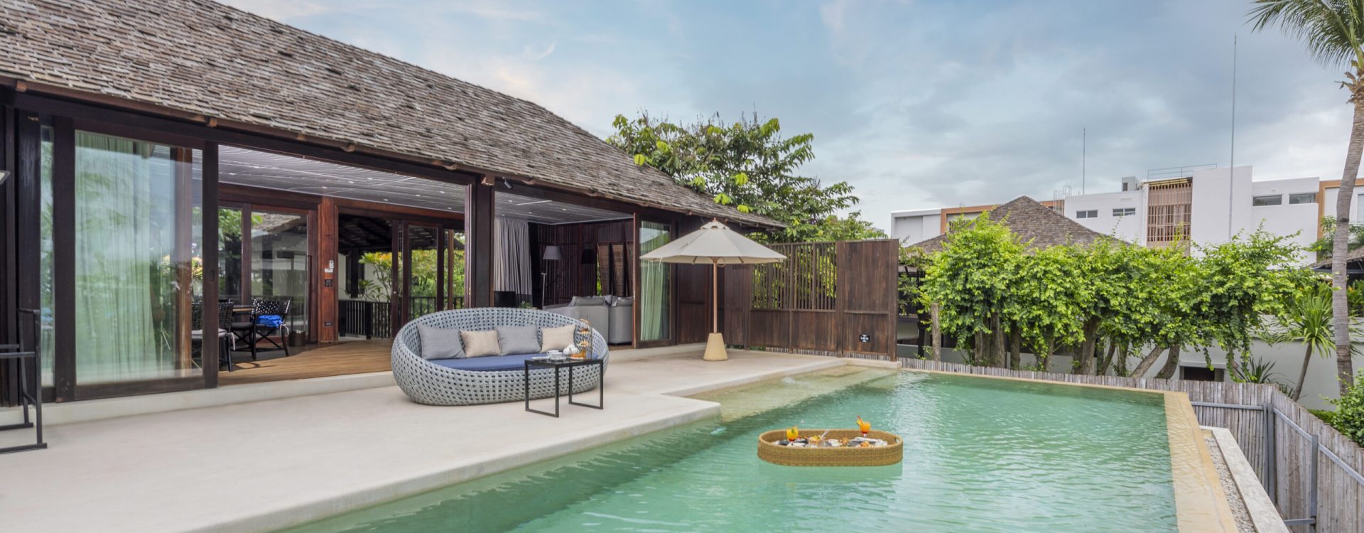 A modern villa with an outdoor pool, lounge chair, circular sofa, and an umbrella. The pool has clear water and a floating tray with drinks and fruit. Lush greenery and blue sky surround the peaceful setting.