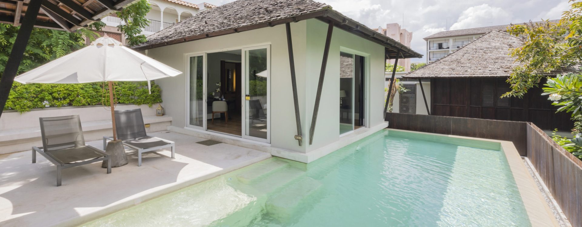 A luxurious villa with a private pool, lounge chairs under an umbrella, and a shaded patio. Glass doors open from the villa to the pool area. Lush greenery and neighboring villas are visible. Bright, sunny weather.