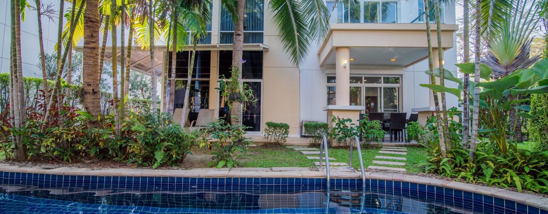 A modern house with large windows, surrounded by palm trees and lush plants, sits behind a blue-tiled swimming pool with metal pool steps. Outdoor patio furniture is visible near the house.