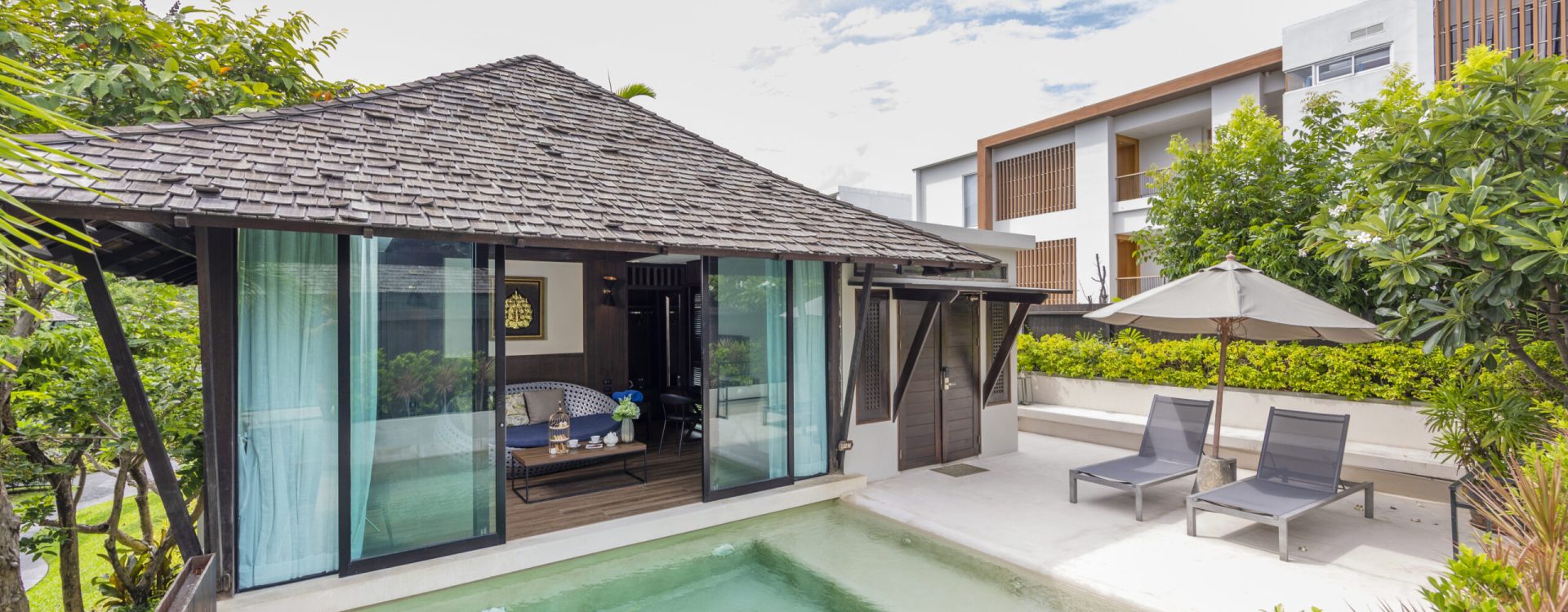 Small modern pool villa with wooden deck, two lounge chairs under an umbrella, and lush greenery surrounding a private swimming pool on a sunny day.