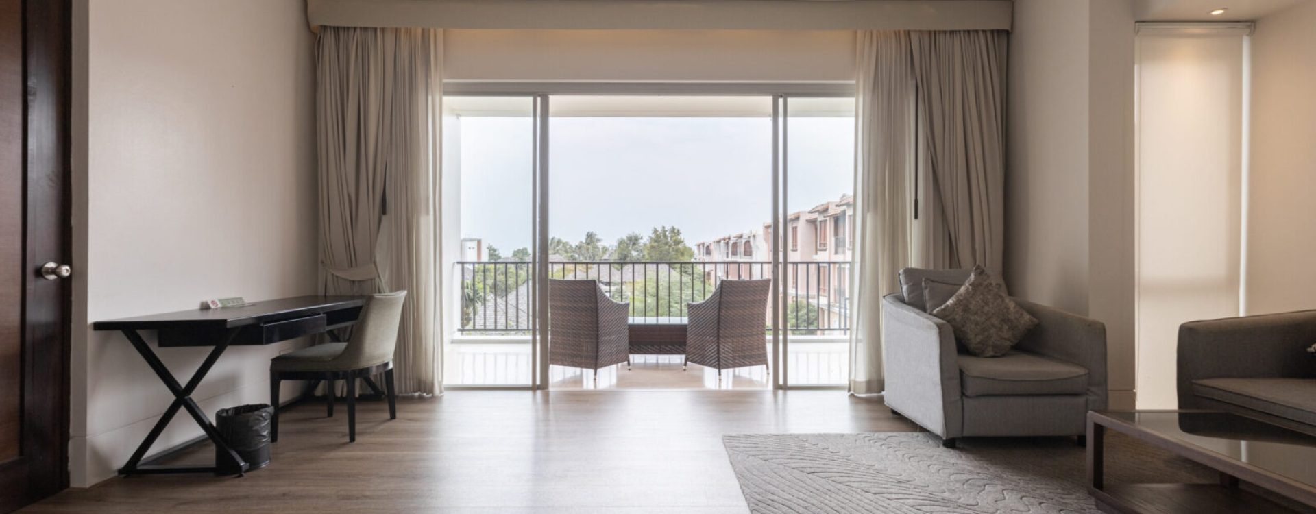 A modern living room with wood floors, a sofa, a desk, and large glass doors leading to a balcony with two chairs. The balcony overlooks trees and neighboring buildings under a cloudy sky.
