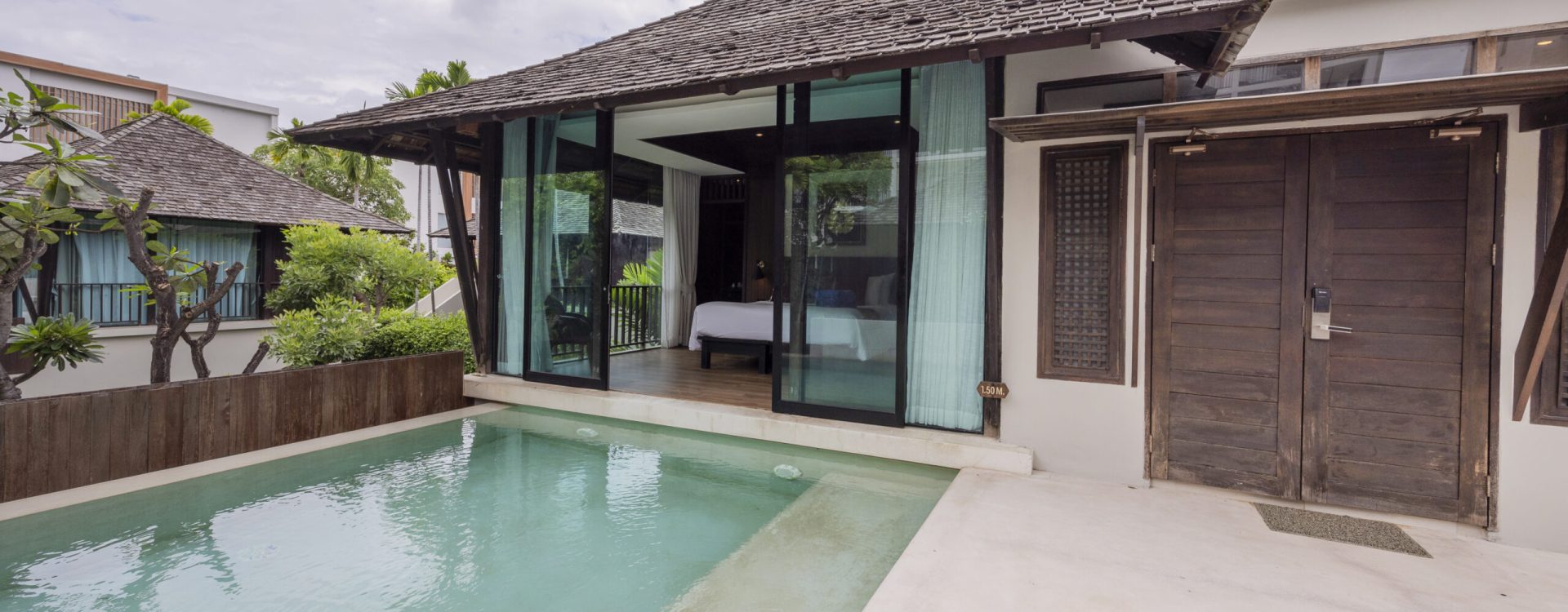 A modern villa with a private pool, glass doors leading to a bedroom, wooden roof, and cloudy sky above. Lush greenery surrounds the peaceful, tropical outdoor area.
