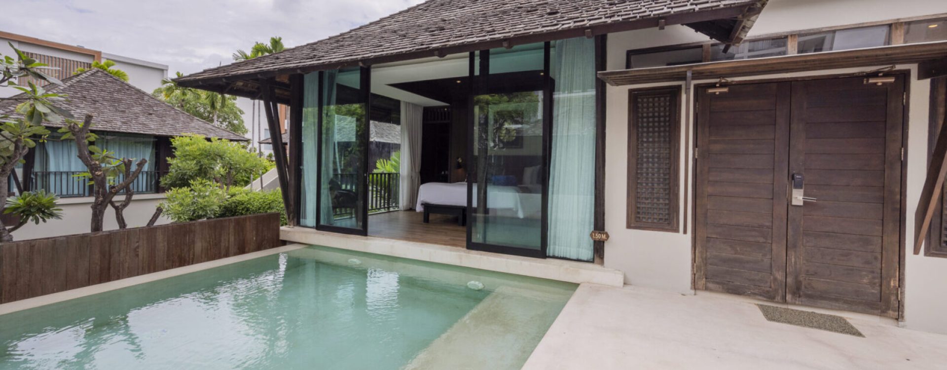 A modern villa with a private pool, glass doors leading to a bedroom, wooden roof, and cloudy sky above. Lush greenery surrounds the peaceful, tropical outdoor area.